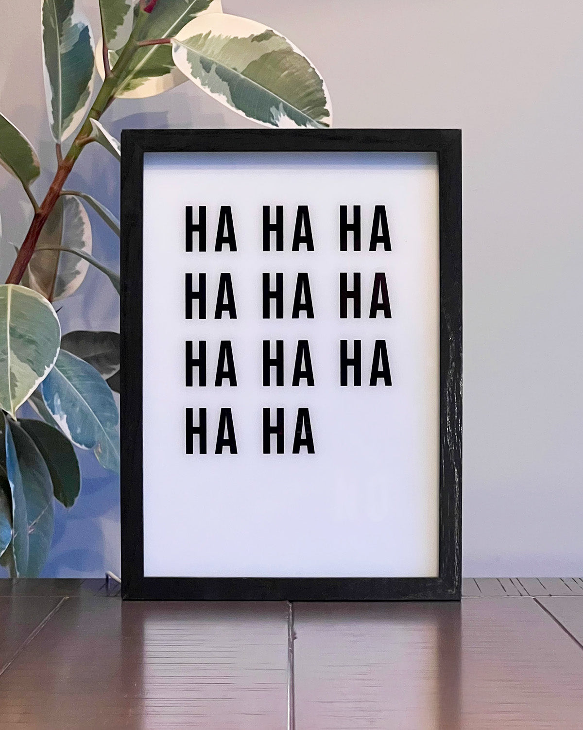 Humorous LED light-up box frame with the quote 'HA HA HA' that transforms to reveal the word 'NO' when lit, set against a modern plant background.